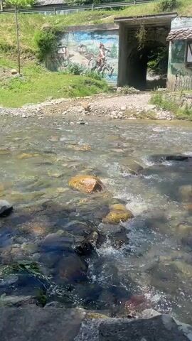 A drone shot pans across a clear, rocky stream. The stream flows through a picturesque village with half-timbered houses and stone walls. A mural depicting a man on horseback is painted on a concrete wall. The drone moves over the water, capturing the sunlight glinting off the surface. The camera then pans to the right, showing more of the village and a lush green hillside. The video ends as the drone continues to move over the stream.