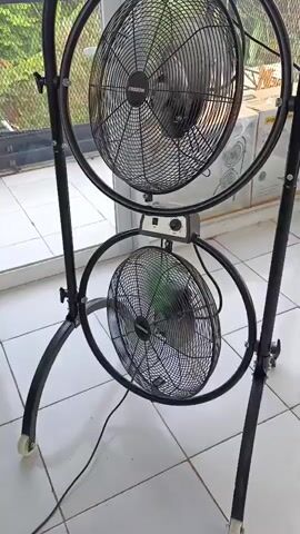 A black metal stand holds two large, round black fans stacked vertically. Both fans have metal grilles and black blades. The stand has curved legs with small wheels at the front. The fans are positioned in front of a window with a view of greenery outside. The floor is tiled with white and grey squares. A power cord is plugged into the lower fan. The video shows a slight wobble in the top fan.