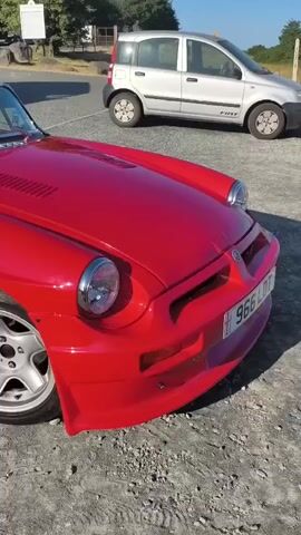 A red convertible sports car is parked on a gravelly surface. The car has its top down and its interior is visible. The seats are black and gray with red trim, and the MG logo is visible on the headrests. The car has a sleek, classic design with prominent headlights and a curved hood. Behind the sports car, a silver Fiat Panda is parked. The background shows trees and a clear blue sky. The overall scene suggests a sunny day in a rural or suburban setting.