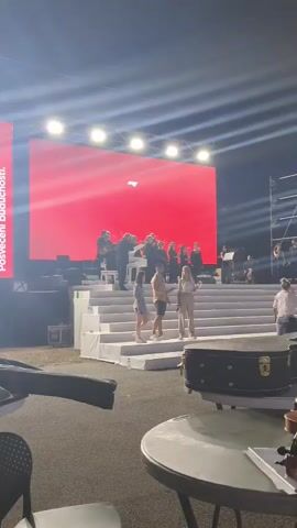 The video shows a wide shot of an outdoor stage at night. A large red screen is visible behind a group of musicians playing instruments. The screen displays the number "35" in white text and the words "U Crnoj Gori" in a script font below it. To the left of the screen, white text reads "Posvećeni budućnosti." Three people are standing on a white staircase in front of the stage. The person on the left is a woman wearing a light-colored denim skirt and a dark top. The person in the middle is a man wearing a light brown t-shirt and shorts. The person on the right is a woman wearing a light-colored pantsuit. The lighting suggests an event or performance. In the foreground, a table and a black instrument case are visible.