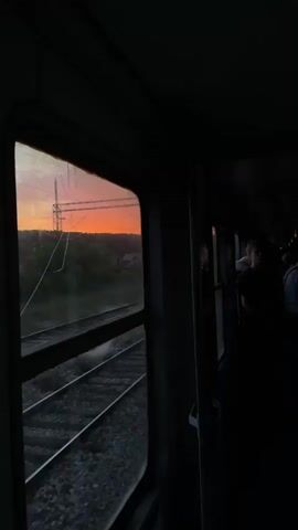 This media shows the view from inside a train car, looking out a window as the sun sets. The sky outside is a vibrant orange and pink, casting a warm glow over the landscape. Silhouetted trees and power lines are visible against the colorful sky. Train tracks stretch into the distance, their gravel beds and metal rails dark against the fading light.

Inside the train car, the lighting is dim, with most of the interior shrouded in shadow. Several people are visible in the background, their faces indistinct in the low light. One person is looking out the window, their profile illuminated by the sunset. The atmosphere feels peaceful and contemplative, perhaps a bit melancholic due to the fading light. The cracked windowpane adds a sense of imperfection and perhaps a hint of the passage of time.

The scene is set in Rakovica Urban Municipality, Serbia, during sunset. The weather appears clear, with no rain or fog evident. The overall impression is one of a journey, with the changing light and the movement of the train suggesting travel and the transition from day to night.