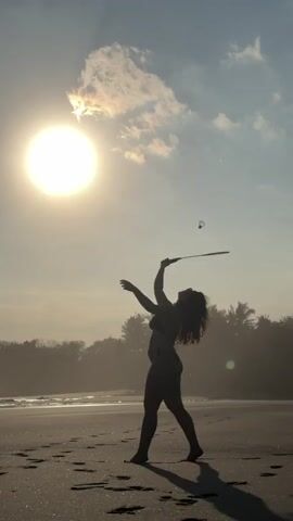 A silhouette of a woman in a bikini is seen on a beach in the evening, facing a bright sun in a hazy sky. She is playing badminton by herself, hitting the shuttlecock into the air multiple times. The camera is static. Palm trees line the horizon behind her.