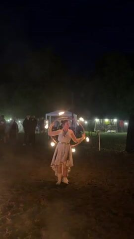 A woman is performing with fire at night. She is wearing a light-colored dress with gold accents and her hair is pulled up. She is holding a hoop with flames around it and appears to be spinning it. She has a headband with markings on her forehead and is wearing gold bracelets on her arms. In the background, there are several people standing and watching, some tents, and a fence. The ground is covered in dirt or sand. There is a fire pit in the foreground, and some trees are visible in the distance. The overall scene suggests an outdoor event or festival at night.