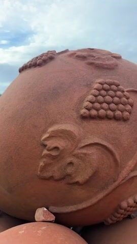 The camera pans out from a large clay pot with an embossed grape design. The pot has a hole in the top. It is sitting on top of a stack of similar clay pots. In the background is a green hillside and a cloudy blue sky.
