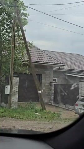 The video shows a view from a car's windshield, which is streaked with water droplets, indicating rain. The camera pans slowly to the right, revealing a residential area. In the foreground, a rough dirt road is visible. To the left of the road, there's a wooden utility pole with some wires and a small white box attached to it. Beyond the pole, there's a tree with green leaves and a brick fence. Further to the right, a house with a brown tiled roof and walls made of grey concrete blocks is visible, with a dark-colored garage door. A wooden fence runs along the property line, separating it from the road. The sky is overcast and gray.
