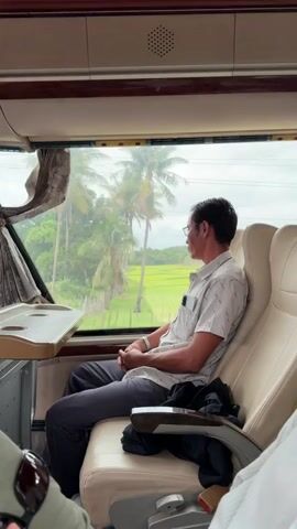A man sits on a bus, looking out the window at a tropical landscape. The bus appears to be in motion, as indicated by the slightly blurred scenery outside. The man is wearing glasses, a light-colored patterned shirt, and dark pants. He is sitting in a tan seat, and there is a tray table attached to the seat in front of him. The view outside the window includes palm trees, green fields, and some buildings. Another person's arm and hand are visible in the foreground, suggesting they are in the seat next to the man. The overall lighting suggests it is daytime, possibly overcast or raining lightly.
