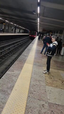 The video shows an underground subway station with a red and white subway train pulling into the station. People are standing on the platform waiting for the train to arrive. The platform is lined with a yellow safety strip with raised dots. The station ceiling is dark and has long, fluorescent lights. Some people are looking at their phones while waiting. The platform is a marble-like material with a speckled pattern.