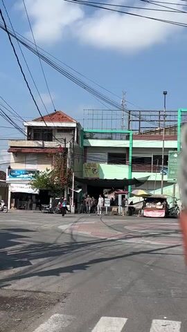 The video captures an outdoor scene on a bright, sunny day with a clear blue sky. In the foreground, a paved road with white pedestrian crossing lines is visible, casting long shadows from overhead wires. A few vehicles, including motorcycles, are parked or in motion. The background features a cluster of buildings, some appearing to be two or three stories high, with visible signage and awnings. One building has a prominent red-tiled roof. Several people are seen walking in front of the buildings and near the road. There are also street vendors with stalls and umbrellas. The overall atmosphere suggests a bustling urban or semi-urban environment.