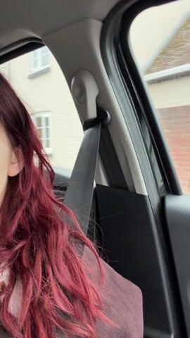 From inside a car, a woman with long, red, wavy hair is seen from the side, with a seatbelt across her shoulder.  Outside the window is a street with houses.  The weather is overcast. The car is moving.  Music is playing.
