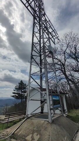 The video shows a view from an elevated vantage point looking out over a landscape. The initial frame shows a metal tower and a small white building. The camera pans right, revealing a blue railing and a sprawling view of green fields and distant mountains under a partly cloudy sky. The camera continues to pan, showcasing more of the landscape and the interplay of clouds and blue sky. The final shot shows a silver railing and green grass in the foreground, with the landscape still visible in the distance.