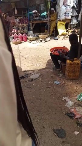 The video shows a bustling outdoor market. There are several stalls with various items for sale, including onions, baskets, and other goods. A young man sits on a yellow container, using his phone while holding a red tray filled with red objects, possibly meat or some sort of snack. The ground is dirty and littered with trash. The camera moves closer to a meat stall where a man in a black t-shirt is cutting large pieces of raw meat. There are several other people around the stall, and someone is paying with a wad of bills.