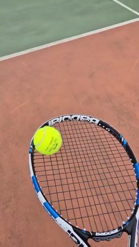 The video shows a tennis racket with a tennis ball balanced on its strings. The racket is white, black, and blue. The tennis ball is yellow. The racket is held in front of a reddish-brown tennis court with white lines. The camera then shifts focus to the tennis ball being hit, rising through the air, passing over the net, and landing on the opposite side of the court. The tennis court is green with white lines. In the background, there are buildings and trees. The sky is overcast.