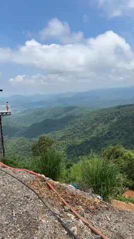 The video shows a panoramic view of lush green rolling hills under a blue sky with scattered white clouds. In the foreground, there is a rocky, uneven slope with some orange dirt and debris, including what appears to be electrical cables. Tall, reedy grass grows along the edge of the slope. The distant hills are layered and appear hazy, suggesting a vast landscape. The overall scene is one of natural beauty and a high vantage point overlooking a verdant, mountainous region. There are no people visible in the video.