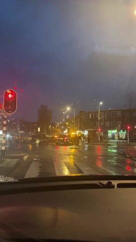 The video shows a view from inside a car at a traffic light at a wet intersection on a dark, rainy evening. The traffic light facing the car is red with a left turn arrow. Other traffic lights at the intersection can be seen in the background. Several cars are stopped at the intersection, including a white car that drives across the intersection. On the right side of the video, several people are standing around a dark-colored car that has its hazard lights flashing. The buildings along the street are brick and appear to be apartments or businesses. The street is wet and reflective, with streetlights adding to the ambient illumination. The sky is a dark, overcast gray.