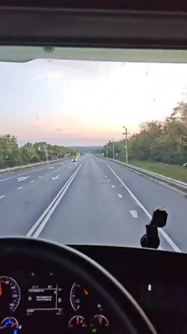 The video shows a first-person view from inside a truck cabin, looking out through the windshield. The truck is on a highway at dusk. The sky is a gradient of orange, pink, and purple. There are trees lining the highway. Other vehicles are visible on the road, including cars and trucks. The truck's dashboard is partially visible, showing speed and other information. The truck appears to be in motion.