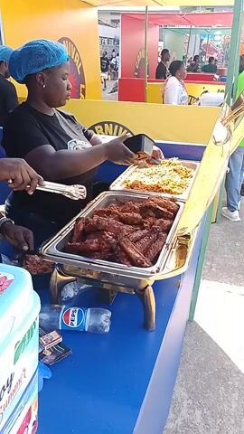 The video shows a food stall at an event. Two people are standing behind a blue counter, serving food from a chafing dish. The chafing dish contains jollof rice and a meat dish. One of the people is wearing a blue hairnet, and the other is wearing a black hat and a red scarf. They are both wearing black t-shirts. There are several cooler boxes labeled "Enjoy Summer" on the counter. The sign above the counter reads "ADUGAN JOLLOF AND FOODS CARNIVAL LAGOS." In the background, there is a large crowd of people.