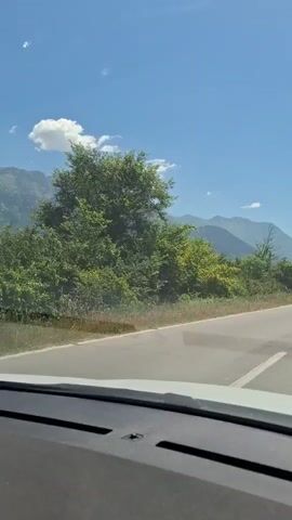 The video shows a view from inside a car driving down a two-lane road on a sunny day. In the opposite lane, a bright red three-wheeled vehicle (likely a motorcycle trike) with two occupants is driving away from the camera. Further down the road, a large, dark vehicle, possibly a military truck, is also heading in the same direction as the trike. The background features lush green trees and shrubs lining the road, with a range of mountains visible in the distance under a clear blue sky with scattered clouds.