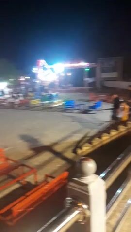 A nighttime aerial shot of an outdoor amusement park. In the foreground, a metal railing with a white post is visible. The camera pans left across a paved area with colorful benches and tables, some occupied by people. Beyond the seating area, a stage with bright lights and a screen is set up. To the right, a set of stairs leads down, with a few people walking down them. The background shows more park attractions, including a Ferris wheel and other rides, illuminated by lights.