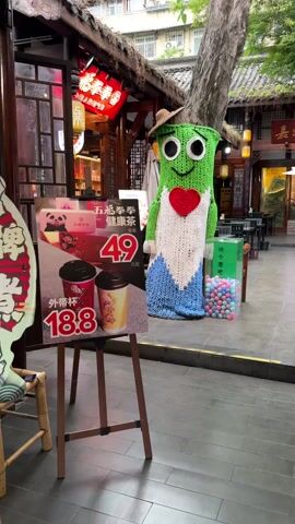 The video shows an outdoor advertisement for a store in what appears to be a market in Asia. A large knitted figure stands to the right, wearing a brown straw hat, with a green head, a white vest with a red heart in the center, and a blue base. Next to it is a clear box filled with pastel-colored balls.  To the left is a sign on an easel advertising tea for 18.8 yuan and a healthy tea set for 49 yuan. The store's sign is in Chinese. The buildings in the background feature traditional Asian architecture. The video is static and doesn't capture any actions or interactions.