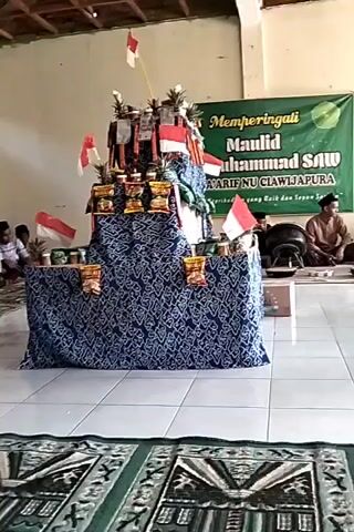 A tiered display, draped in dark blue fabric with a white patterned design, sits in the center of a room. The display is decorated with Indonesian flags, small bags of snacks, and what appear to be pineapples. A banner with green and white text hangs on the wall behind the display. To the right of the display, a man in traditional clothing is seated on the floor. The room has tiled flooring and what appears to be a wooden ceiling. In the foreground, a patterned rug is visible. The overall scene suggests a celebration or event, possibly religious, given the banner and the man's attire.