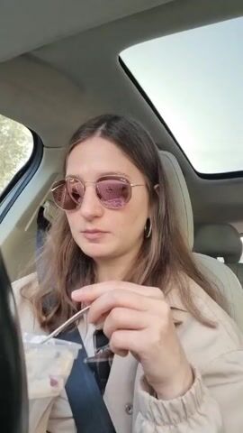 A woman wearing a beige jacket and rose-tinted aviator sunglasses is sitting in the driver's seat of a car with a sunroof. She holds a clear plastic container and a plastic spoon, eating a meal from the container. She looks at the camera while she eats. The car's interior is visible with the steering wheel on the left and the passenger seat on the right. Trees are visible outside the car.