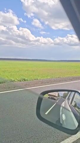 The video shows a view from inside a car looking out the window and into the side mirror. The car is on a paved road with a white line. To the left of the road is a large, flat green field with some scattered vegetation. In the distance, a dark line of trees marks the horizon. Above the field, the sky is blue with fluffy white clouds. The side mirror reflects the road behind the car, showing other vehicles and the interior of the car. The camera perspective suggests the video is being recorded from a moving vehicle.