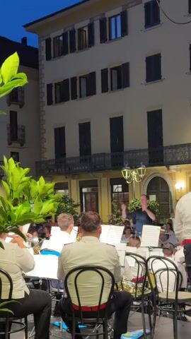 The video shows an outdoor concert at dusk. A conductor, dressed in a dark shirt, stands on a platform directing a brass band. The band members, some wearing white shirts with red trim, are seated and playing instruments. They are positioned in the foreground, with music stands and chairs visible. Behind the band, a building with multiple windows and a balcony is illuminated. The sky is dark blue, suggesting it is evening. An audience is visible in the background, seated and standing, watching the performance. Lush green foliage is in the left foreground, partially obscuring the view. The lighting appears to be a mix of natural twilight and artificial stage lighting.