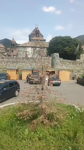 The video captures an exterior view of a historic stone building, possibly a monastery, with a domed roof and arched doorways. In the foreground, there is a dry, bare tree. Several vehicles are parked in a lot in front of the building, including dark-colored cars and a white van. There are also small wooden stalls set up in the parking lot. A few individuals are visible near the stalls and the cars, with some appearing to interact with each other or load items into vehicles. The area is surrounded by stone walls and lush green hills in the background, under a bright, partly cloudy sky.