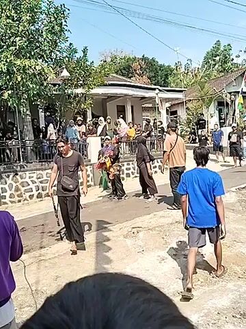 A group of people are gathered in an outdoor street setting, likely for a cultural performance or event. In the foreground, a person wearing a purple hoodie is partially visible, facing away from the camera. To their right, a person in a blue t-shirt and shorts walks away from the viewer. A group of onlookers are positioned on a raised porch-like area behind a low stone wall. Several individuals are standing and observing a performance taking place in the street.

In the street area, a male performer wearing a black t-shirt and dark pants is holding a long, thin object, possibly a prop weapon. To his right, a performer in traditional attire, including a headdress and colorful clothing, is in motion, possibly dancing or performing. Another individual in a black outfit and hijab is also present. Further back, a drummer is visible behind a sound system. The environment appears to be a rural or residential area with houses and trees. The sky is bright and clear, suggesting a daytime event. The overall atmosphere suggests a lively gathering and a cultural display.