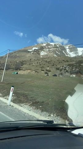 A car is driving on a paved road through a mountainous area with patches of snow on the hillsides. The sky is blue with a few white clouds. Power lines run along the side of the road. There are some buildings scattered in the landscape. The car is moving forward, and the view is from the driver's perspective.