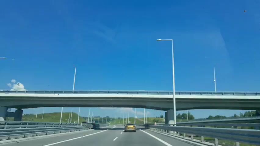 The video shows a POV shot from a vehicle driving on a highway under a clear blue sky. The road has multiple lanes, with a yellow car in front and other vehicles in the distance. There are guardrails on the sides and a bridge overhead. Tall lampposts line the highway. The surrounding landscape consists of green hills and some scattered clouds.