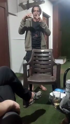 Here is a description of the video:

The video shows a man standing in front of a chair while talking and gesturing with his hands. He is wearing a green hoodie, a black t-shirt, and dark shorts. He is barefoot. There are two other people in the video, seated around a small table. On the table are cans of Budweiser beer, a glass of beer, and other items. The setting appears to be an outdoor patio or covered area with artificial turf flooring. A door and a section of white wall are visible in the background. There is a light fixture above the door, casting a purple beam.