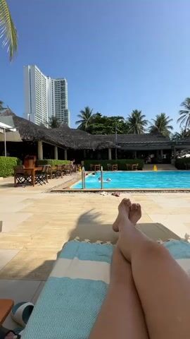 A person's legs are in the foreground, crossed at the ankles, resting on a light blue and white striped towel on a lounge chair. The legs are tanned. Behind the person is a tiled patio with lounge chairs and umbrellas. To the left, there is a thatched roof building with tables and chairs. Further back, a bright blue swimming pool is visible, with several people swimming. Beyond the pool and the thatched roof building, palm trees are abundant. To the far left and center background, a tall, white skyscraper stands against a clear blue sky. The sun is shining, casting shadows on the patio.