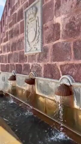 The video shows a long, stone fountain with three spouts, set into a red brick wall.  Each spout is shaped like a half-dome or mushroom, with water flowing continuously from each, accumulating in the trough below.  A stone carving with a heraldic crest and crown is mounted above the center of the fountain.