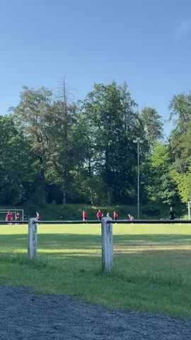 The video shows a soccer field with a group of people playing soccer. The people are wearing red and black jerseys. There are trees in the background and a clear blue sky. The video is taken from a low angle, looking up at the field. The overall scene is a peaceful outdoor activity.