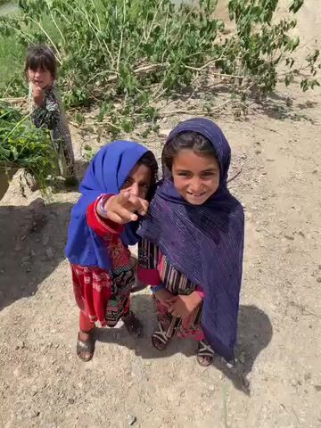 This outdoor image, captured in Ashoqah, Afghanistan, features three young girls under bright daylight. In the foreground, two girls are the main subjects, looking towards the camera.

The girl on the left is wearing a blue headscarf and a red and black patterned dress with long sleeves. Her right arm is raised, and she is pointing directly towards the viewer with her index finger. Her face is partially obscured by her hand and the headscarf, but she appears engaged. She also wears multiple colorful bangles on her right wrist.

Beside her, on the right, another girl is prominently featured, smiling broadly at the camera. She is dressed in a dark blue or purple patterned headscarf that covers her hair and shoulders, along with a striped dress that has pink accents. Both girls are wearing open-toed sandals.

In the background on the far left, a third girl is partially visible, holding a bundle of green leafy plants or branches. She has dark hair and is looking towards the left, away from the camera.

The setting appears rural, with a dry, light-colored dirt and gravel ground. Behind the girls, there are green trees and bushes, suggesting a natural environment. The clear lighting indicates a sunny day, possibly mid-morning or afternoon, given the visible but not harsh shadows. The overall mood conveyed by the girls' expressions is one of curiosity and friendliness, particularly from the smiling girl in the foreground. No text is visible in the image.