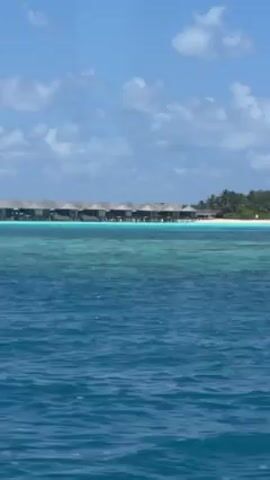 The video shows a row of overwater bungalows at a tropical resort. The bungalows have a similar architectural style, with dark roofs and light-colored sides, and are situated on stilts above the turquoise water. Behind the bungalows is a white sandy beach, lined with palm trees and other vegetation. The sky is blue with scattered clouds. The water in the foreground is a deeper blue and has small waves. The video appears to be shot from a moving boat, facing the resort.