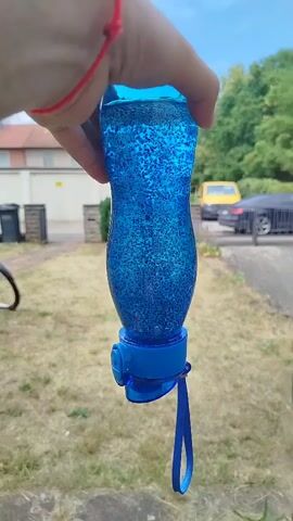A hand with a red string bracelet is holding up a blue plastic water bottle filled with a blue liquid and glitter. The bottle has a blue cap with a blue strap attached. The background shows a blurry outdoor scene with dry grass, a fence, trash bins, a bicycle wheel, a yellow van, and a black car. The lighting suggests it is daytime.