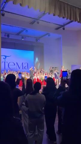 A group of performers is on stage taking a final bow at the end of a performance. The performers are a mix of children and adults. Most of the children wear red and white costumes in the style of traditional folk dress, while others, and the adults, wear more formal attire like dresses and blazers. Many people in the audience are recording the moment on their smartphones. The stage has a large screen displaying the word "Тема" in Cyrillic script. Stage lighting illuminates the performers.