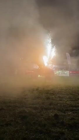 A person in dark clothing with a long skirt is standing in a grassy area at night, holding a large flamethrower that emits bursts of fire into the air. A dark vehicle, possibly a tractor, decorated with Christmas lights and producing white sparks from its top, is positioned behind the person to the right. The scene is illuminated by the fire, lights, and sparks, with thick smoke or fog filling the air. Several people are visible in the background, dimly lit. The ground is covered in short, dark grass.