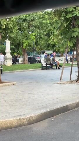 A video captures a street scene with a park area. In the foreground, a gray sidewalk is visible, bordered by a curb. Beyond the sidewalk, a paved walkway extends into a park area with scattered trees. A white bench sits in the park area, occupied by two men facing away from the camera. Both men are casually dressed. Behind the bench, more trees and some parked vehicles are visible, including a black car and a white van. A white pillar is on the far left. The overall atmosphere appears to be an outdoor public space.