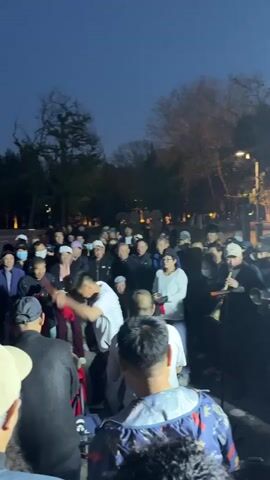 Here is a description of the video:
A crowd of approximately 50 people, primarily adults wearing hats, gathers in what appears to be a park at dusk. Several individuals are playing musical instruments, including drums, cymbals, and a brass instrument. The drum player dances while playing, eliciting cheers and applause from the crowd. Trees and streetlights are visible in the background.