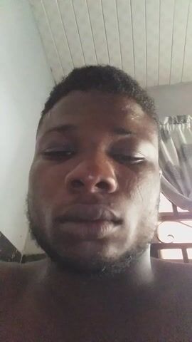 A close-up shot shows the face of a Black man. The camera angle is looking up at him. He has short, dark hair and a beard. His eyes are partially closed. The background includes a white ceiling and what appears to be a window with white blinds. The man's upper body is visible, suggesting he may be shirtless. A voice speaks briefly.
