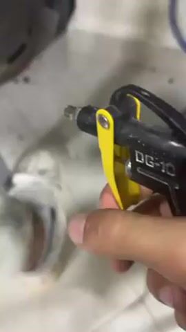 A hand is holding a black air blow gun with a yellow trigger, identified as a DG-10 model, and is activating it to blow air onto a blurred object on a light gray surface.