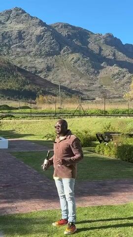 A man stands on a brick patio holding a bottle of wine, gazing towards mountains in the background. He wears a brown shirt, light jeans, and sunglasses. The setting includes a green lawn, a wire fence, and glimpses of trees and vegetation against a clear blue sky.