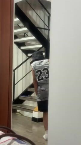 A person wearing a gray Jordan jersey, black shorts, a black bucket hat, and white socks, with the brim of the hat covering their face, dances in a mirror. The person moves their arms, head, and legs, appearing to dance. Behind the person in the mirror is a wooden wardrobe and a staircase with white steps and black railings. Part of a white wall is visible to the right of the person. A blurred pillow with blue and white designs is visible in the bottom left corner.