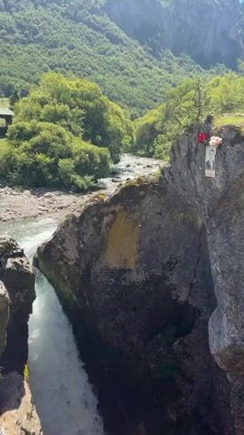 A video shows a rushing river cascading down a rocky gorge. On the edge of the cliff overlooking the gorge, a framed portrait of a man is attached to the rock face, along with some red flowers. The surrounding landscape is lush and green, with trees covering the mountainside. The sky is blue and clear.