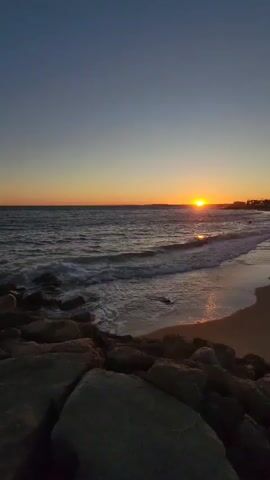 The video shows a sunset at a beach. The sky is clear and a deep blue at the top, fading to a warm orange near the horizon where the sun is setting. The sun casts a golden reflection on the ocean water, which has gentle waves rolling onto a sandy beach. There are rocks in the foreground, some of which are wet from the waves. In the distance, along the coastline, there are some buildings and silhouetted figures of people. The overall atmosphere is peaceful and serene.