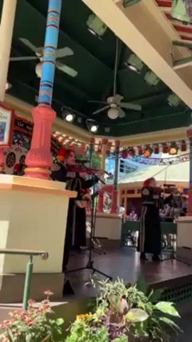 Three mariachi musicians are performing on a small stage within a gazebo-like structure. The musicians, all women, are dressed in traditional black mariachi suits adorned with gold trim. Two of them are playing violins, while the third plays a stringed instrument, possibly a guitarron. Each has a microphone in front of them. The gazebo is decorated with colorful pennants and has a dark green ceiling with several ceiling fans. The stage is bordered by a low fence with planters filled with greenery. People can be seen seated at tables in the background. The scene is well-lit, suggesting it is daytime.