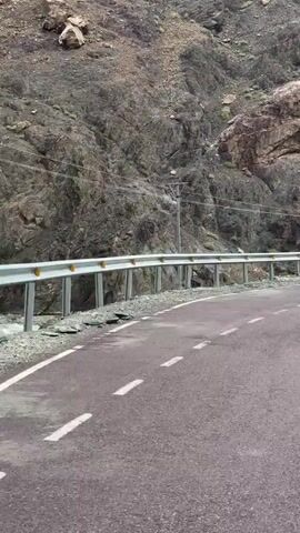 The video shows a road alongside a steep, rocky mountain. A metal guardrail runs along the edge of the road, separating it from the mountain. In the middle of the video, three people are visible near the guardrail, along with a bicycle. One person, wearing a blue jacket and a grey hood, is pointing towards the mountain. Another person, wearing a red jacket, is standing next to them. A third person, wearing a blue jacket, is partially visible. The road appears to be paved and has white dashed lines marking the lanes. The environment is mountainous and appears to be in a remote or natural area.